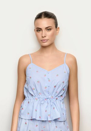 Kobieta ubrana w jasnoniebieski, paskowany, bezrękawnik z małymi różowymi kwiatowymi wzorami i wiązaniem w talii, stojąca na jednolitym tle.
