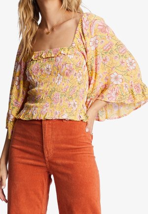 Kvinde iført gul blomstrede smock-bluse med flæset firkantet udskæring og brede klokkeærmer, kombineret med højtaljede rustrøde kordbuks.