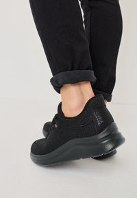 Black athletic shoes with a textured fabric upper, lace-up design, and a thick rubber sole. Ankle-height with slight curve at the heel.