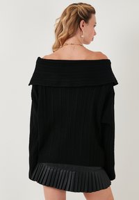 Suéter negro acanalado con hombros al aire y un cuello ancho. La modelo lo lleva sobre una minifalda corta de polipiel negra con pliegues.