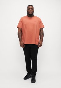Korte mouwen, zachte terracotta-kleurige t-shirt met klein logo op de borst, gecombineerd met zwarte slim-fit broek en zwarte sneakers.