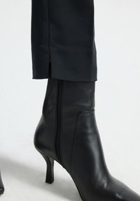 Bottines en cuir noir à talon effilé, avec une fermeture éclair sur le côté et une texture lisse, assorties à un pantalon sombre à coupes ajustées.