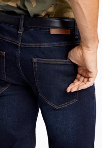 Jeans de mezclilla oscura con costuras naranjas, con bolsillos traseros. Una mano se está metiendo en el bolsillo trasero derecho. Cinturón negro visible en la parte superior.