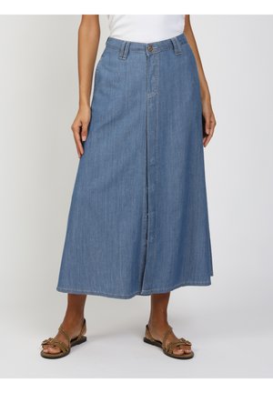 Frau trägt einen blauen denim A-Linien-Midirock mit Vorderverschluss, kombiniert mit braunen flachen Sandalen mit Nieten und einem weißen Oberteil.