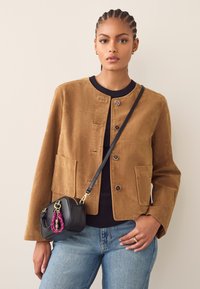 Bruine corduroyjas met een ronde hals, voorzakken en zilveren knopen. Gecombineerd met een blauwe spijkerbroek en een kleine zwarte tas met een roze accent.