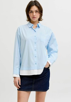 Nuori nainen, jolla on vaaleansininen napillinen paita valkoisen topin päällä ja tumma farkkuhame, seisoo yksiväristä taustaa vasten.