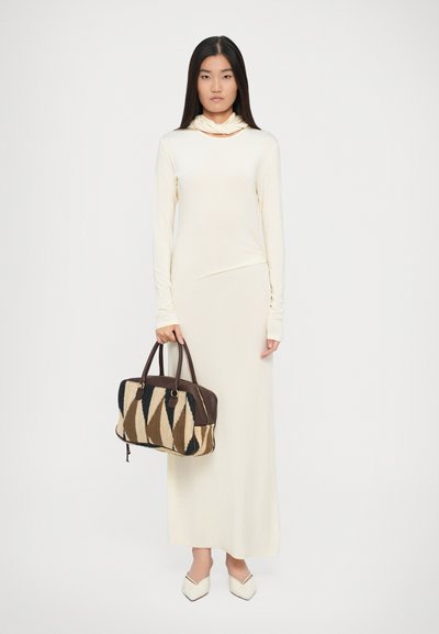 Femme dans une longue robe crème tenant un grand sac à main à motifs marron et noir, portant des chaussures crème à bouts pointus, se tenant devant un fond uni.