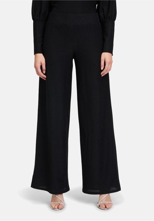 Zwarte, wijde broek gemaakt van een zachte, gestructureerde stof. Voorzien van een elastische tailleband en een vloerlength ontwerp. Duidelijke details van de schoenen zichtbaar.