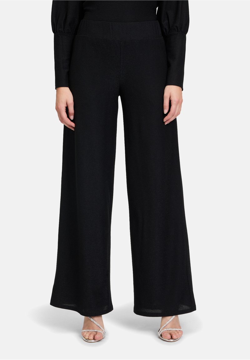 Zwarte, wijde broek gemaakt van een zachte, gestructureerde stof. Voorzien van een elastische tailleband en een vloerlength ontwerp. Duidelijke details van de schoenen zichtbaar.