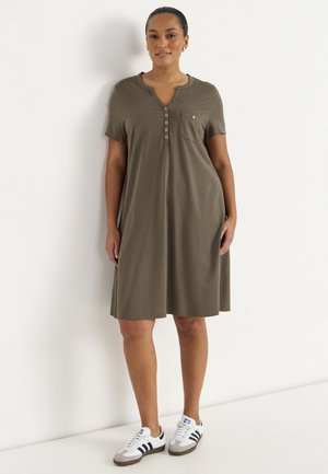 Kvinde iført kort, olivengrøn kjole med knapper og brystlomme samt hvide Adidas-sneakers, stående mod en hvid væg.