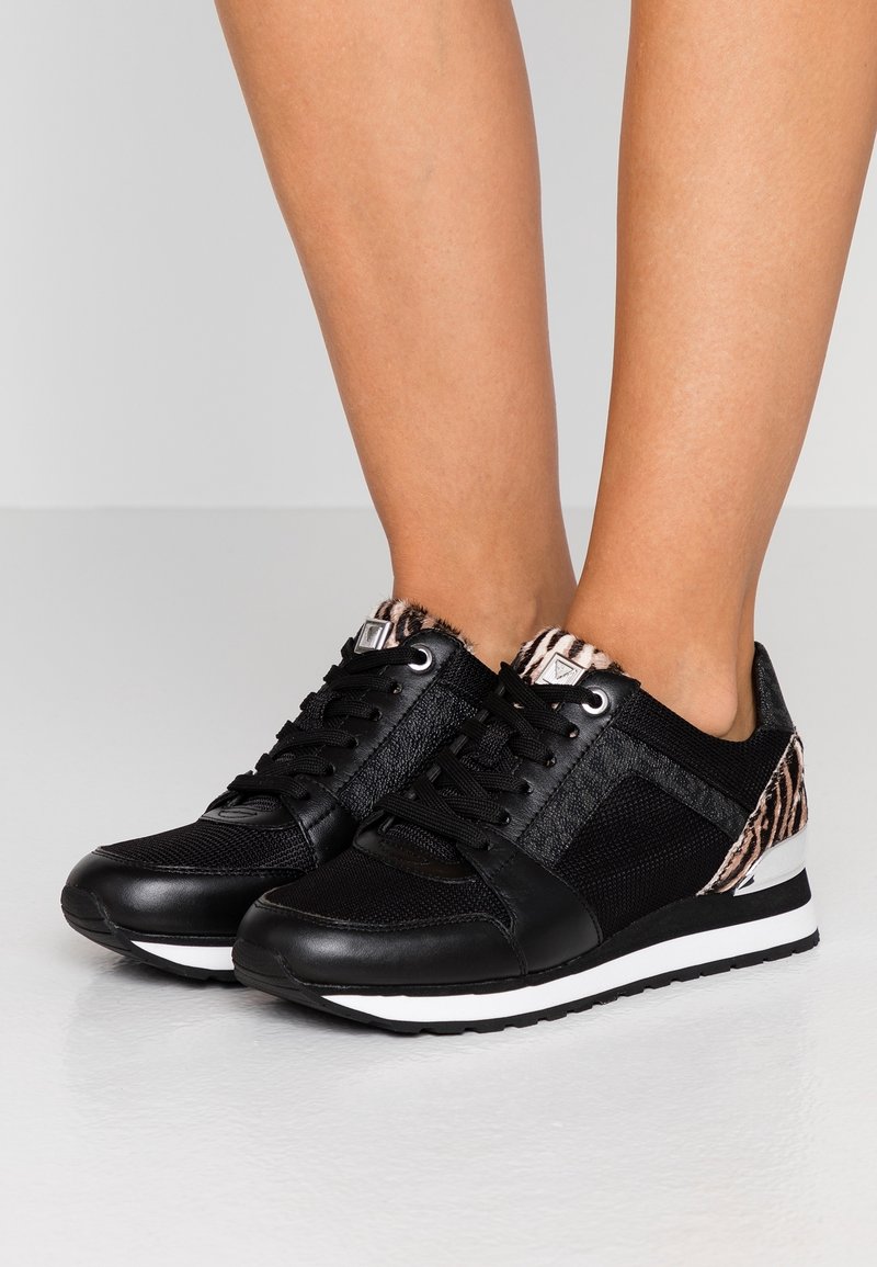 Baskets noires à lacets avec semelles blanches, présentant des détails en maille et en cuir, des accents à imprimé léopard sur le talon et la languette, portées sur des pieds nus.