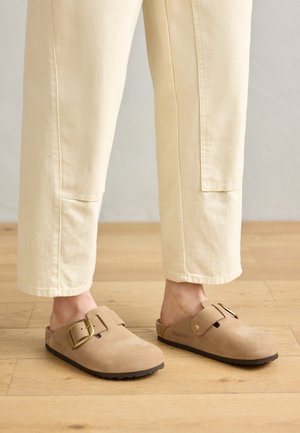 Hnědé semišové dřeváky s širokým páskem a zlatou přezkou, texturovaným povrchem a hnědou gumovou podrážkou; nošeny s volnými krémovými tapered kalhotami.