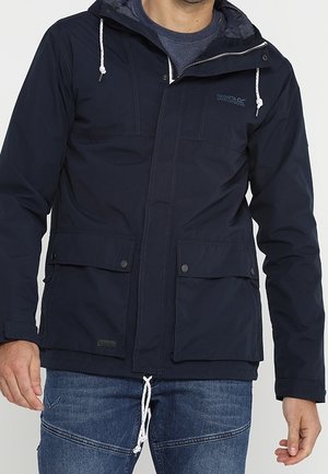 Homme portant une veste à capuche bleu marine avec poches avant et fermeture éclair, associée à un jean bleu sur un fond uni.