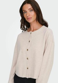 Jasnoróżowy krótki kardigan wykonany z miękkiego materiału, z okrągłym dekoltem i pięcioma brązowymi guzikami z przodu. Długie rękawy o luźnym kroju.
