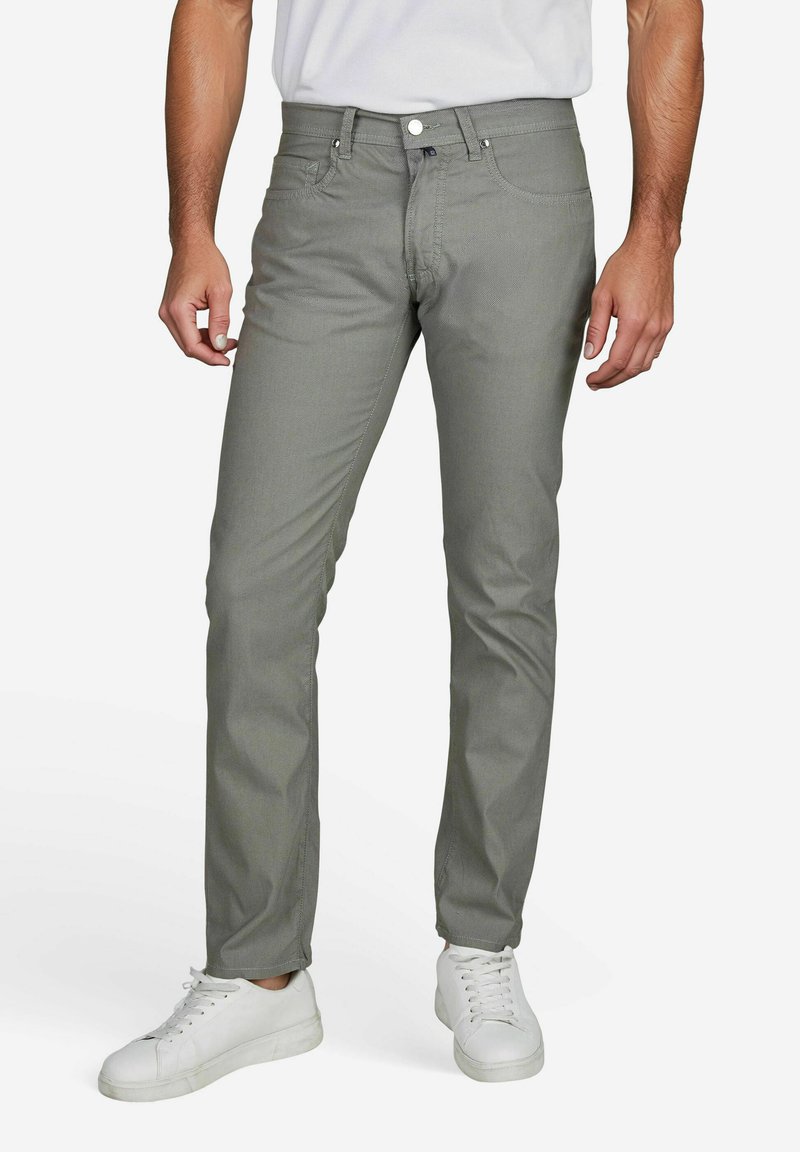 Man draagt strakke grijze broek met witte sneakers en een wit overhemd dat in de broek is gestopt, staand tegen een effen witte achtergrond.