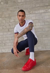 Rote Sneakers mit strukturierten Seitenpaneelen, getragen mit navy Leggings und einem weißen Shirt mit einem gerippten roten und blauen Kragen, auf einem strukturierten Boden.