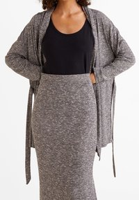 Grauer gestrickter langer Cardigan mit gebundenen Ärmeln, über einem schwarzen Tanktop und einem passenden grau-taillierten Rock. Strukturierter Stoff überall.
