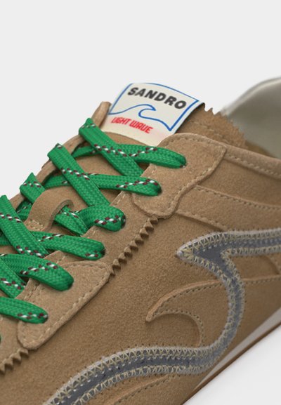 Κρεμ σουέντ αθλητικό παπούτσι με πράσινα κορδόνια, λεπτομέρειες ραφής σε ζιγκ-ζαγκ και ετικέτα "SANDRO LIGHT WAVE" στη γλώσσα.