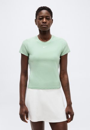 Kvinde med kort hår iført en lysegrøn tætsiddende Nike t-shirt og hvid nederdel, stående mod en ensfarvet baggrund.