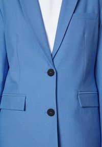 Jasnoniebieska marynarka o gładkiej fakturze, zapinana na jeden czarny guzik, z dwiema przednimi kieszeniami i klapami w kształcie ucha.