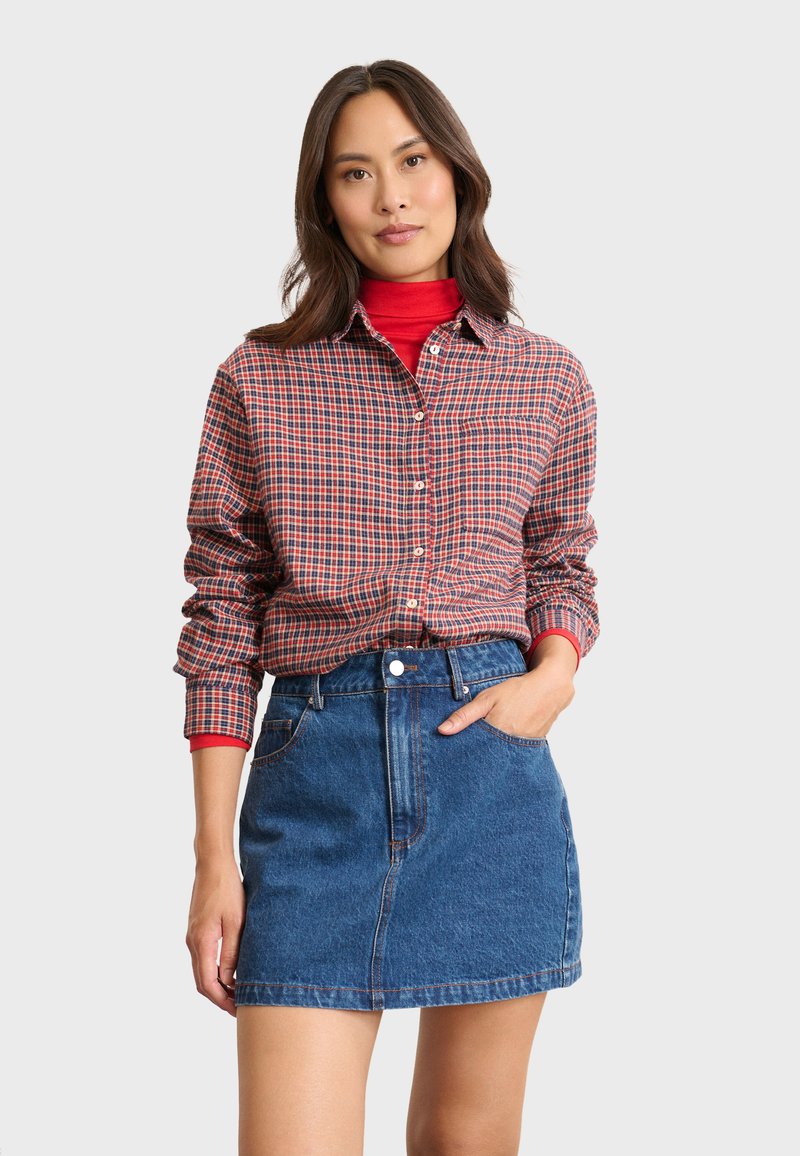 Chemise à carreaux rouge et marine à boutons avec des manches longues, portée sur un col roulé rouge. Associée à une mini-jupe en denim bleu avec des poches.