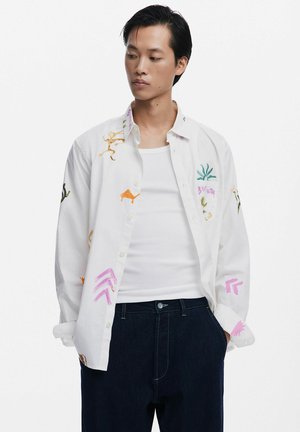 Weißes Hemd mit Knöpfen und bunten gestickten Designs, darunter Tiere und Pflanzen, kombiniert mit einem schlichten weißen Tanktop und dunklen Jeans.