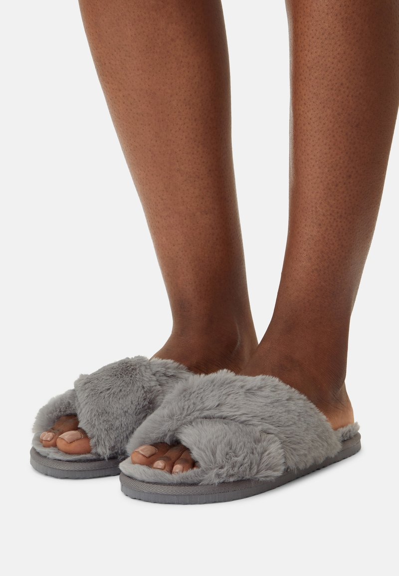 Pieds portant des chaussons gris pelucheux à bouts ouverts avec des brides croisées sur un fond blanc uni.