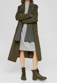 Manteau long vert olive avec larges revers, robe en maille grise, et bottines marron à lacets. Caractérisé par un tissu texturé et un look superposé.
