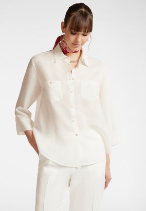 White, long-sleeve, button-up shirt in lightweight fabric with two chest pockets and pearly buttons; paired with white trousers.