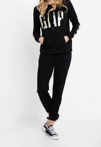 Sudadera negra con cremallera y "GAP" en letras doradas reflectantes, combinada con pantalones joggers negros a juego y zapatillas negras con suelas blancas.