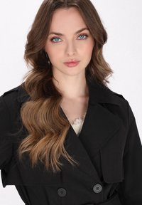 Fekete trench kabát dupla gombolású kivitelezéssel, széles hajtókákkal és kiemelkedő gombokkal. A modellen hosszú, hullámos haj és diszkrét smink látható.