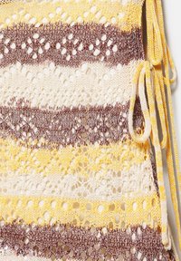 Tissu tricoté avec des rayures en dentelle ajourée jaunes, brunes et crème, avec des cordons de fil jaune et marron noués sur le côté.