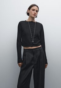 Svart kort ärmad cardigan med knappstängning, tillverkad av ett mjukt material. Kombineras med högmidjade mörka byxor som har en avslappnad passform.