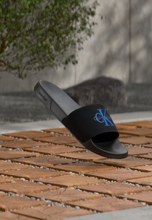Sandalia tipo slide negra de Calvin Klein con logo CK azul en la tira, flotando sobre un suelo de madera y baldosas de piedra al aire libre.
