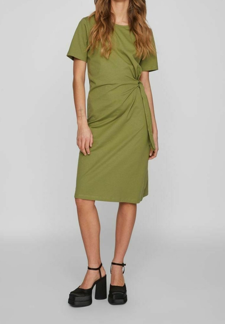 Femme portant une robe vert olive longueur genou avec des manches courtes et un nœud côté, associée à des talons compensés noirs avec brides à la cheville.
