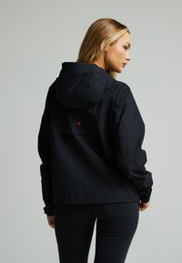Zwarte waterdichte jas met een capuchon en elastische manchetten, met een klein rood logo op de achterkant. Glad textuur en losse pasvorm.