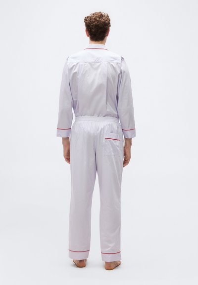 Άνδρας στέκεται ξυπόλυτος, γυρισμένος με την πλάτη, φοράει λευκή πυτζάμα με μακριά μανίκια, κόκκινη επένδυση και τσέπη στην πίσω δεξιά πλευρά.