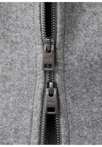Szary materiał z mieszanki wełny o teksturowanej powierzchni, wyposażony w podwójny zamek błyskawiczny z metalowymi elementami i logo MNG.