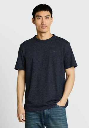 Mężczyzna w granatowym, teksturowanym t-shircie z krótkim rękawem i niebieskich dżinsach, stojący z jedną ręką w kieszeni na jasnym tle.