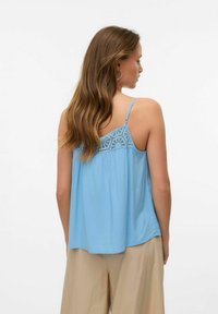 Jasnoniebieski top na ramiączkach z koronkowym detalem z tyłu, gładki materiał, cienkie regulowane ramiączka i luźny krój, połączony z beżowymi spodniami.