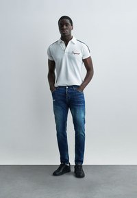 Homme adulte debout les mains dans les poches, portant un polo blanc, un jean bleu et des baskets noires sur un fond gris uni.