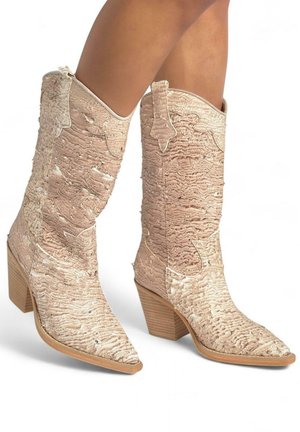 Botas de tobillo de estilo occidental en beige claro con material texturizado, punta afilada y tacón de madera apilada. Presenta acentos de costura decorativa.