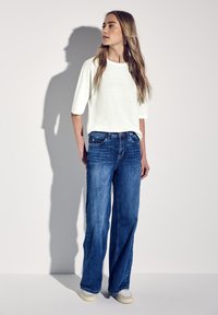 Hvid, løstsiddende, kortærmet top parret med blå vide jeans. Modellen bærer lyse sneakers og viser en afslappet, uformel stil.