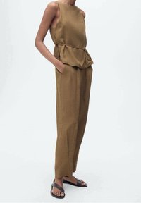 Combinaison sans manches marron avec taille peplum, poches latérales et jambes fuselées. Fabriquée dans un tissu texturé, associée à des sandales noires.