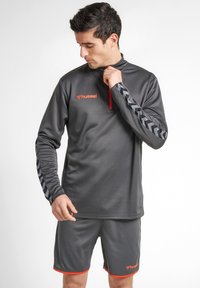 Grå sportstopp med hög krage och halvdragkedja, med texturerat tyg och röda detaljer, i kombination med matchande shorts och logodetaljer.