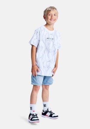 T-shirt graphique blanc avec motif de flammes, logo noir, associé à un short en denim clair et des baskets noires, blanches et rouges.