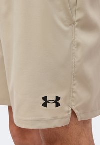Μπεζ αθλητικό σορτς με μαύρο λογότυπο Under Armour κοντά στο τελείωμα, φορεμένο στο πάνω μέρος του μηρού ενός ατόμου.