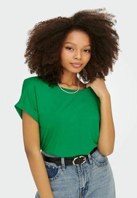 Fiatal nő dús, göndör hajjal, zöld pólót, gyöngy nyakláncot, fekete övet és magas derekú kék farmert viselve, fehér háttér előtt áll.