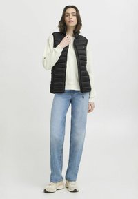 Gilet rembourré noir avec un col montant, fermeture éclair et coutures horizontales. Assorti à un sweat léger et un jean bleu clair.