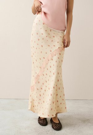 Frau trägt einen cremefarbenen floralen Maxirock mit rosa Spitzenbesatz, eine rosa Satinbluse und dunkelbraune gewebte Ballerinas, steht auf einem hellen Boden.